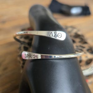May include: A silver-colored cuff bracelet with the Greek letters &Delta;&Sigma;&Theta; engraved on one end and "SPR '26" on the other. The bracelet is curved and open, designed to be worn on the wrist. The background is blurred, with a wooden surface and a patterned fabric visible.