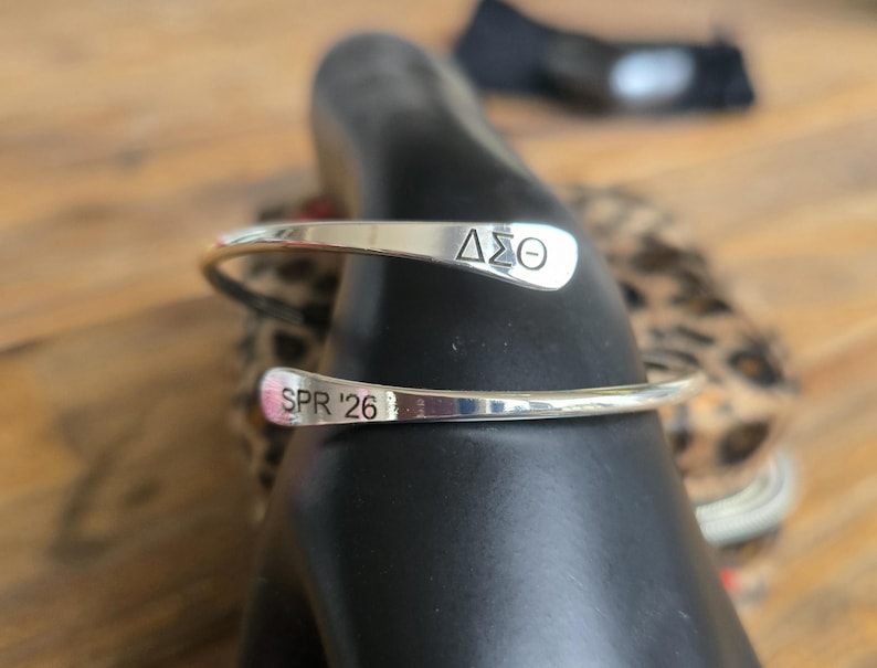May include: A silver-colored cuff bracelet with the Greek letters &Delta;&Sigma;&Theta; engraved on one end and "SPR '26" on the other. The bracelet is curved and open, designed to be worn on the wrist. The background is blurred, with a wooden surface and a patterned fabric visible.