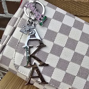 May include: A silver-toned metal keychain featuring the letters AKA in a vertical arrangement, a charm with the number 25, and a pink and green charm with the text SKEE-WEE, all attached to a white and grey checkered handbag with a red edge.