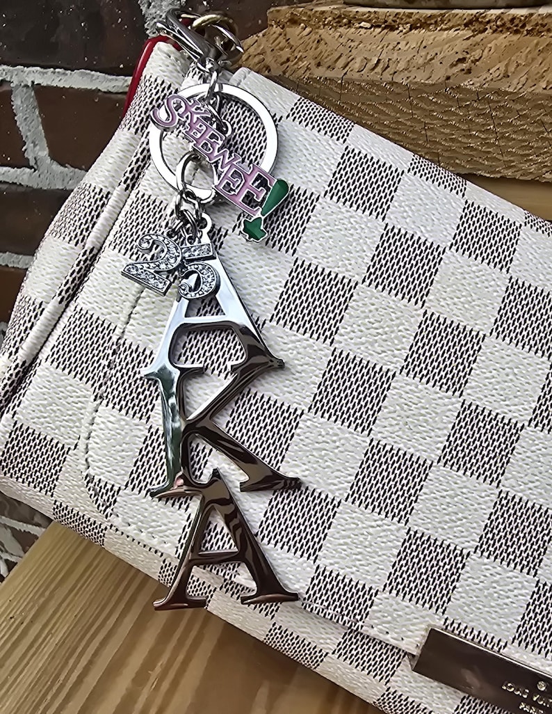 May include: A silver-toned metal keychain featuring the letters AKA in a vertical arrangement, a charm with the number 25, and a pink and green charm with the text SKEE-WEE, all attached to a white and grey checkered handbag with a red edge.