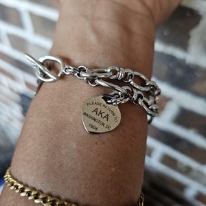 May include: A silver-toned toggle bracelet featuring a heart-shaped charm engraved with the text PLEASE RETURN TO AKA WASHINGTON, DC 1908. A second gold-toned curb chain bracelet is worn below it on the wrist, showcasing a classic link design.