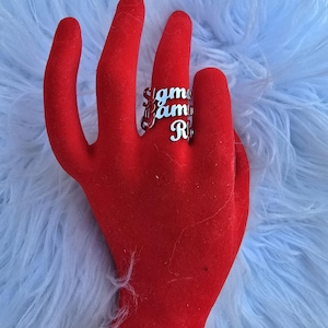 May include: Silver ring with the words "Sigma Fam" and "R.I.P." stacked on top of each other. The ring is on a red velvet hand model.