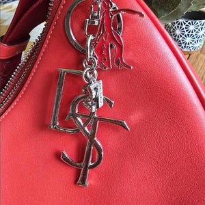 May include: A bright red handbag featuring a silver-toned metal keychain attachment. The keychain consists of a silver ring, a red and silver figure charm, a large silver letter D, a silver YSL logo, and a small silver number 7 charm.