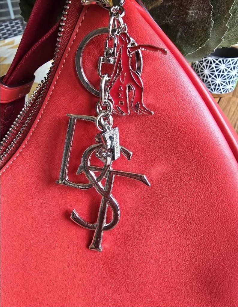 May include: A bright red handbag featuring a silver-toned metal keychain attachment. The keychain consists of a silver ring, a red and silver figure charm, a large silver letter D, a silver YSL logo, and a small silver number 7 charm.