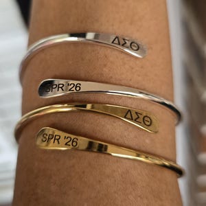 May include: A close-up of three open cuff bracelets. Two are silver, and one is gold. Each bracelet has engraved text: two say "SPR '26" and two say "&Delta;&Sigma;&Theta;". The bracelets are worn on a person's arm.