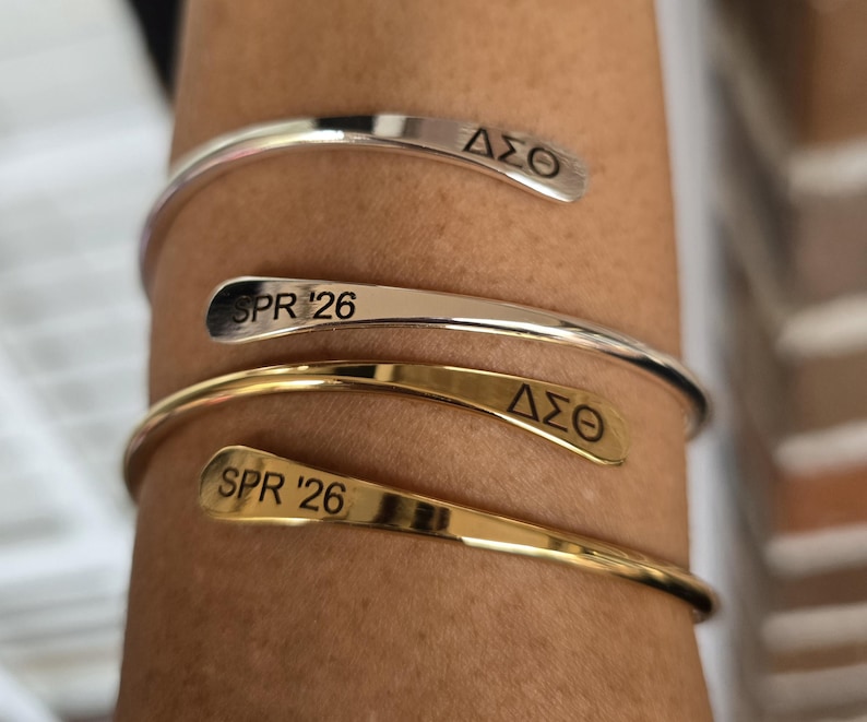 May include: A close-up of three open cuff bracelets. Two are silver, and one is gold. Each bracelet has engraved text: two say "SPR '26" and two say "&Delta;&Sigma;&Theta;". The bracelets are worn on a person's arm.