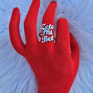 May include: A silver ring with the words "Zeta Phi Beta" engraved on it. The ring is worn on a red velvet hand.
