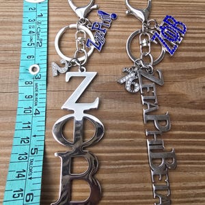 May include: Two silver-toned metal keychains with lobster clasps. One features the Greek letters Z&Phi;B and a number 2 charm, the other reads ZETAPHIBETA with a 1920 charm. A blue measuring tape shows the items are approximately 6.5 inches long.