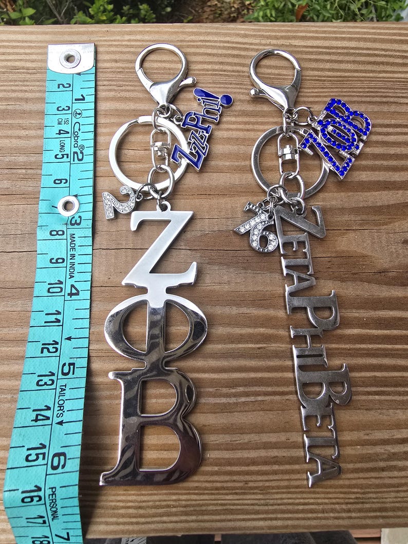 May include: Two silver-toned metal keychains with lobster clasps. One features the Greek letters Z&Phi;B and a number 2 charm, the other reads ZETAPHIBETA with a 1920 charm. A blue measuring tape shows the items are approximately 6.5 inches long.