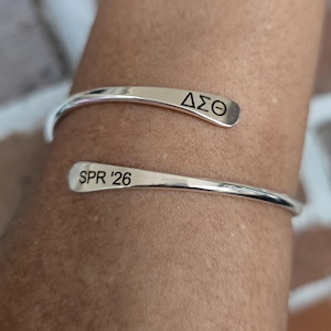 May include: A silver cuff bracelet with the Greek letters &Delta;&Sigma;&Theta; and the text "SPR '26" engraved on the ends. The bracelet is open and wraps around a person's wrist, showcasing a minimalist design.