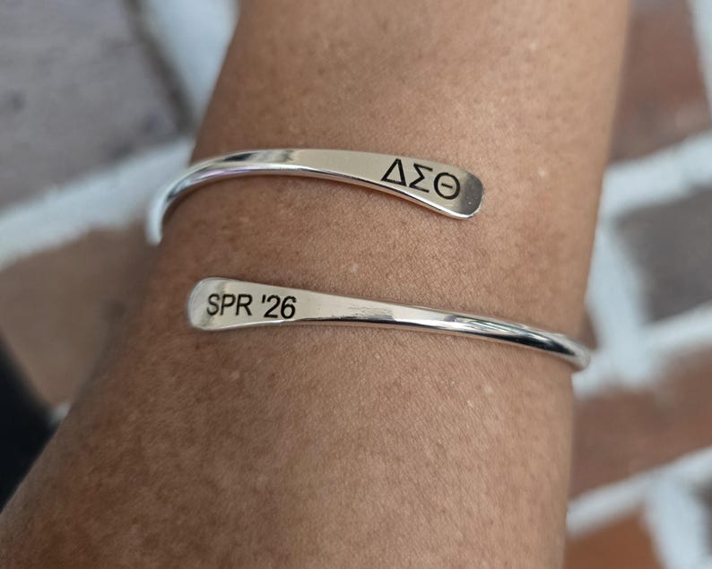 May include: A silver cuff bracelet with the Greek letters &Delta;&Sigma;&Theta; and the text "SPR '26" engraved on the ends. The bracelet is open and wraps around a person's wrist, showcasing a minimalist design.