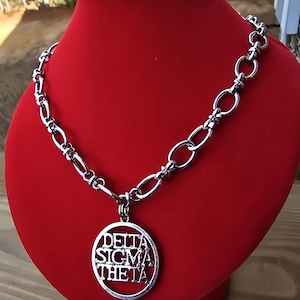 May include: A silver chain necklace with a round pendant that says "Delta Sigma Theta" in a silver font.