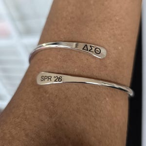 May include: A silver-colored cuff bracelet with the Greek letters &Delta;&Sigma;&Theta; engraved on one end and "SPR '26" on the other. The bracelet is wrapped around a person's arm.