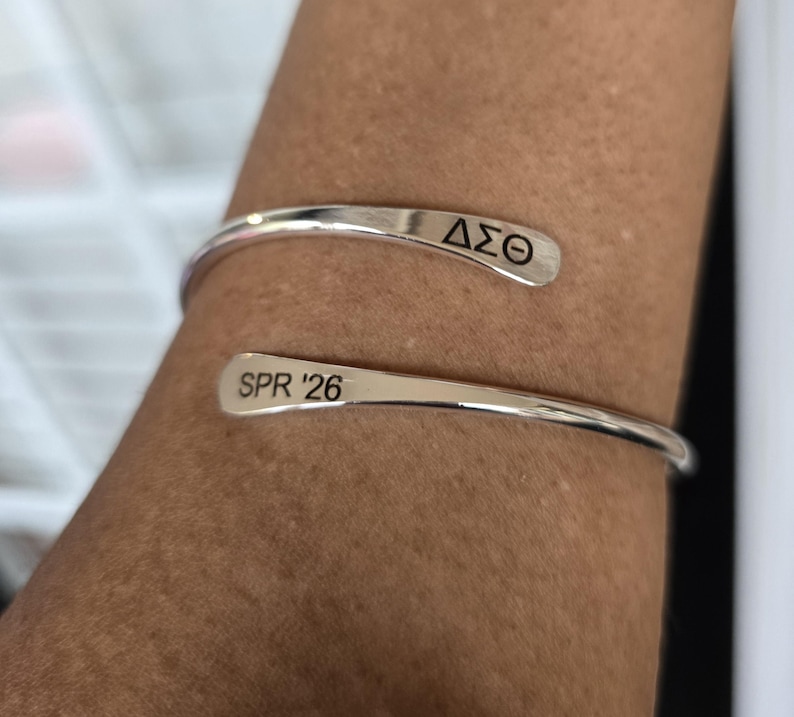 May include: A silver-colored cuff bracelet with the Greek letters &Delta;&Sigma;&Theta; engraved on one end and "SPR '26" on the other. The bracelet is wrapped around a person's arm.
