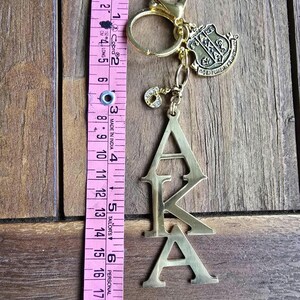 May include: A gold-toned metal keychain featuring the letters AKA stacked vertically, a shield-shaped charm, and a small rhinestone-encrusted number 6 charm. A pink measuring tape placed alongside shows the keychain length is approximately 6 inches.