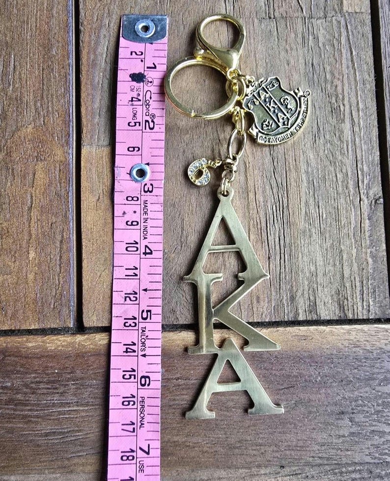 May include: A gold-toned metal keychain featuring the letters AKA stacked vertically, a shield-shaped charm, and a small rhinestone-encrusted number 6 charm. A pink measuring tape placed alongside shows the keychain length is approximately 6 inches.