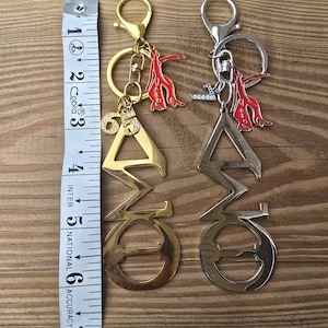 May include: Two metal keychains featuring Greek letters, one gold-toned and one silver-toned, each with a red figure charm and a rhinestone-accented number 65 or letter L. A white measuring tape shows the keychains are approximately 6.5 inches long.