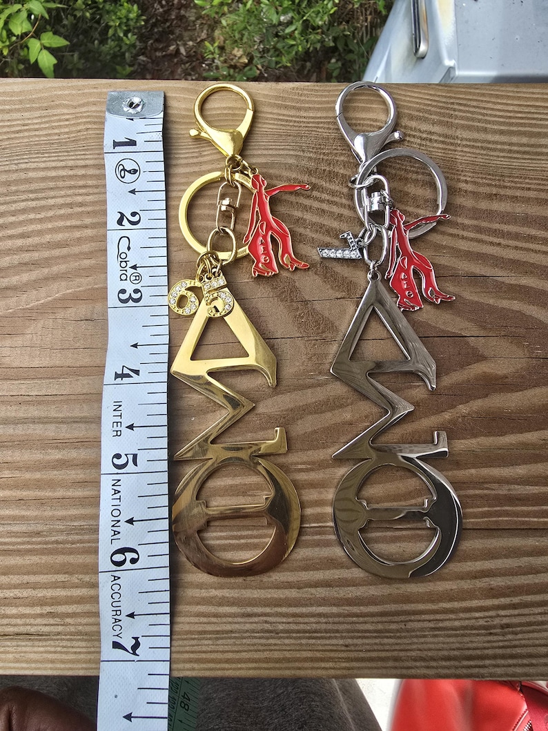 May include: Two metal keychains featuring Greek letters, one gold-toned and one silver-toned, each with a red figure charm and a rhinestone-accented number 65 or letter L. A white measuring tape shows the keychains are approximately 6.5 inches long.