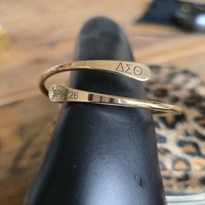 May include: A gold-colored cuff bracelet with the Greek letters &Delta;&Sigma;&Theta; and the text SPR 26 engraved on it. The bracelet is wrapped around a black object, with a blurred background.