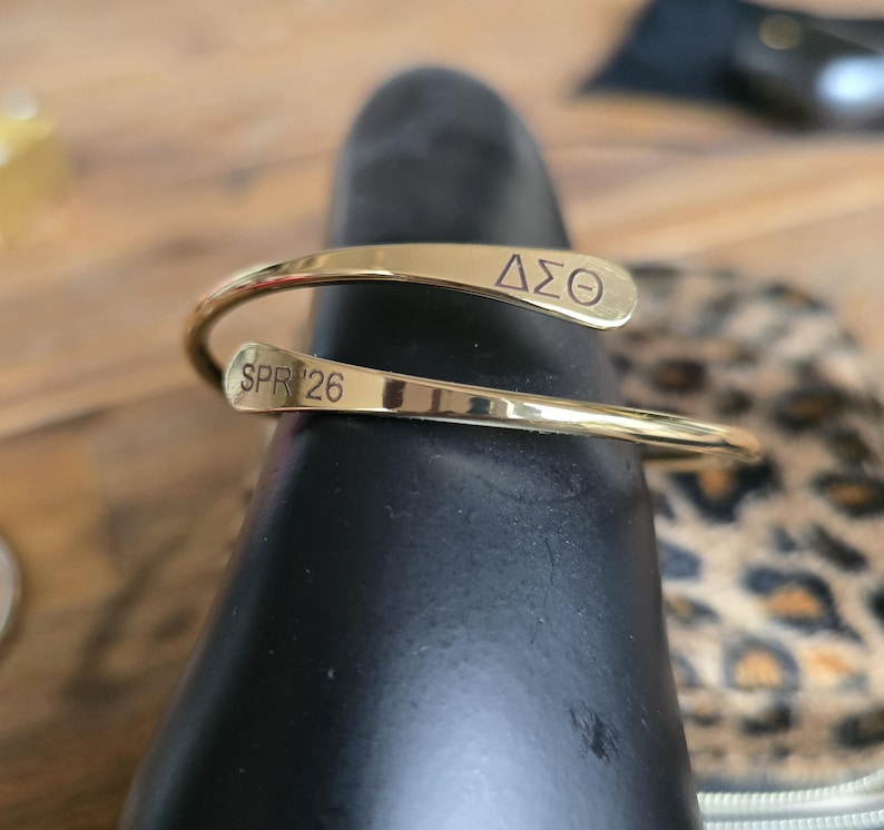 May include: A gold-colored cuff bracelet with the Greek letters &Delta;&Sigma;&Theta; and the text SPR 26 engraved on it. The bracelet is wrapped around a black object, with a blurred background.