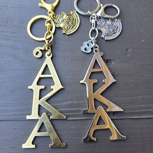 May include: Two metal keychains featuring the letters AKA in a vertical arrangement. One keychain is gold-toned with a number 6 charm, and the other is silver-toned with a number 8 charm. Both include a small shield-shaped emblem with Greek letters.
