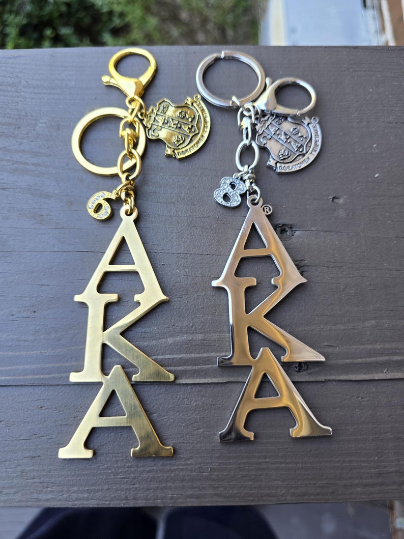 May include: Two metal keychains featuring the letters AKA in a vertical arrangement. One keychain is gold-toned with a number 6 charm, and the other is silver-toned with a number 8 charm. Both include a small shield-shaped emblem with Greek letters.
