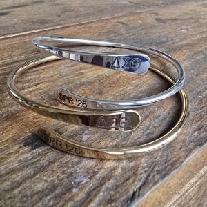 May include: Two metal wrap bracelets, one silver and one gold, rest on a wooden surface. Each bracelet features a curved design with engraved text and a Greek letter symbol. The silver bracelet has "&Delta;&Sigma;&Theta;" and "SPR '26" engraved.