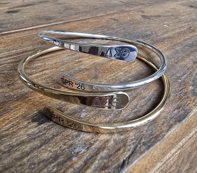 May include: Two metal wrap bracelets, one silver and one gold, rest on a wooden surface. Each bracelet features a curved design with engraved text and a Greek letter symbol. The silver bracelet has "&Delta;&Sigma;&Theta;" and "SPR '26" engraved.