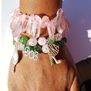 May include: A set of four bracelets featuring pink and green crystal beads, a charm with the text "1908", a charm with the text "AKA", and a charm shaped like a high heel shoe.