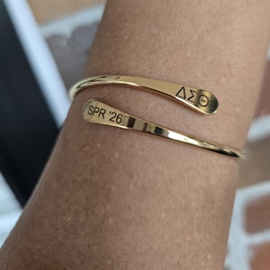 May include: A gold-colored cuff bracelet with the engraved text "SPR '26" and the Greek letters "&Delta;&Sigma;&Theta;". The bracelet has a curved, open design. The image shows the bracelet on a person's arm.