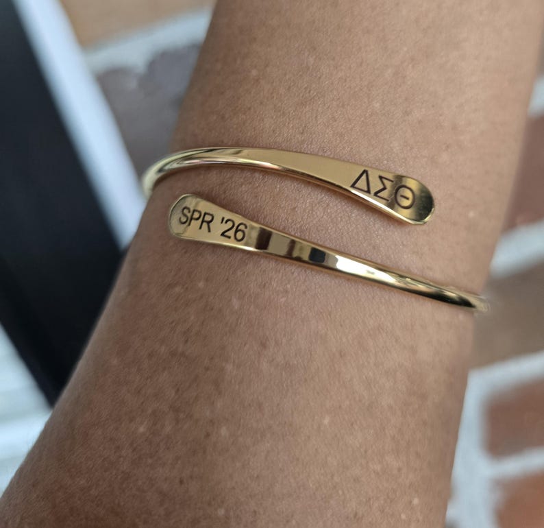May include: A gold-colored cuff bracelet with the engraved text "SPR '26" and the Greek letters "&Delta;&Sigma;&Theta;". The bracelet has a curved, open design. The image shows the bracelet on a person's arm.