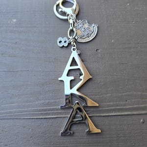 May include: A silver-toned metal keychain featuring the letters AKA in a vertical arrangement. Attached to the split ring are two charms: one is a shield-shaped crest with intricate detailing, and the other is a small number eight encrusted with clear stones.