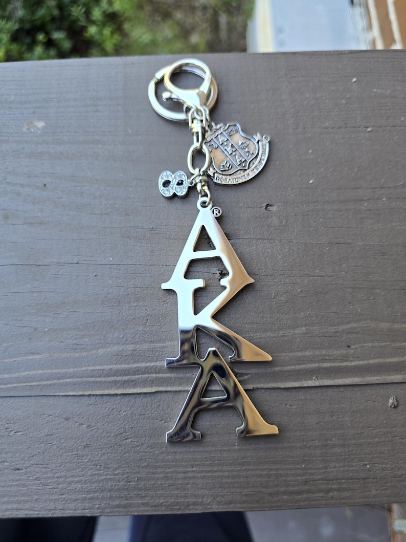 May include: A silver-toned metal keychain featuring the letters AKA in a vertical arrangement. Attached to the split ring are two charms: one is a shield-shaped crest with intricate detailing, and the other is a small number eight encrusted with clear stones.