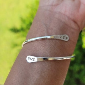 May include: A silver-toned bypass cuff bracelet featuring a smooth, polished finish. One end of the bracelet is engraved with the Greek letters ΣΓΡ, while the opposite end is engraved with the number 1922 in a clean, black sans-serif font.