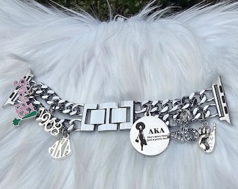 Puede incluir: Una correa de reloj de metal plateado con un diseño de cadena de eslabones dobles y múltiples dijes. Los dijes incluyen el texto 1908, AKA, la silueta de una persona con afro, el texto She's more than just a pretty face y un dije de gesto manual.