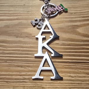 May include: A silver-toned metal keychain featuring the letters AKA stacked vertically. The charm includes a small pendant with the number 25 in rhinestones, a pink and silver charm with the text SOROR, and a small green and silver leaf-shaped charm.