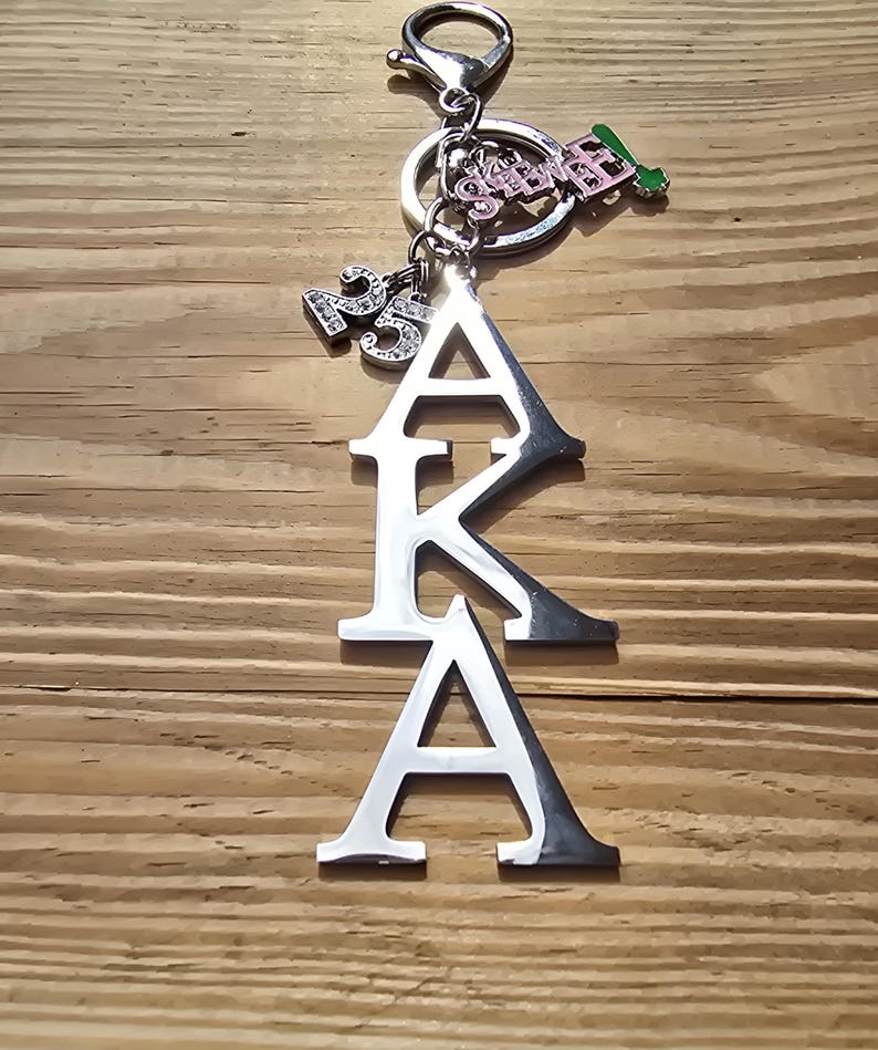 May include: A silver-toned metal keychain featuring the letters AKA stacked vertically. The charm includes a small pendant with the number 25 in rhinestones, a pink and silver charm with the text SOROR, and a small green and silver leaf-shaped charm.