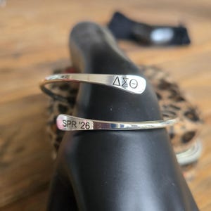 May include: A silver cuff bracelet with the Greek letters &Delta;&Sigma;&Theta; engraved on one end and "SPR '26" on the other. The bracelet is displayed on a black form, with a blurred background.