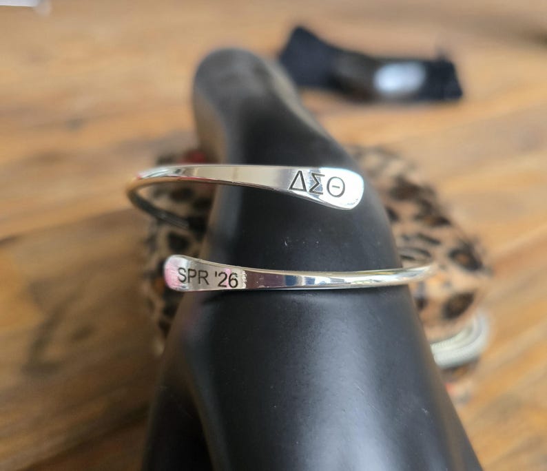 May include: A silver cuff bracelet with the Greek letters &Delta;&Sigma;&Theta; engraved on one end and "SPR '26" on the other. The bracelet is displayed on a black form, with a blurred background.