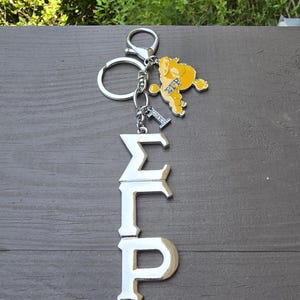 May include: A silver-toned metal keychain featuring the Greek letters Sigma Gamma Rho in a vertical arrangement. Attached to the split ring is a yellow poodle charm with the letters &Sigma;&Gamma;&Rho; in blue, and a small silver charm shaped like the number one.