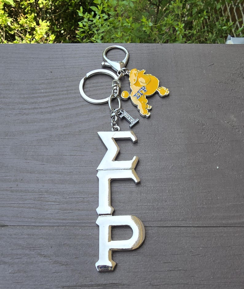 May include: A silver-toned metal keychain featuring the Greek letters Sigma Gamma Rho in a vertical arrangement. Attached to the split ring is a yellow poodle charm with the letters &Sigma;&Gamma;&Rho; in blue, and a small silver charm shaped like the number one.