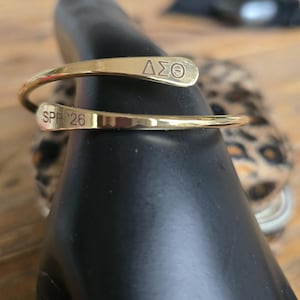 May include: A gold-colored cuff bracelet with the Greek letters &Delta;&Sigma;&Theta; and the text "SPR '26" engraved on it. The bracelet is wrapped around a black object, with a blurred background featuring a leopard print.