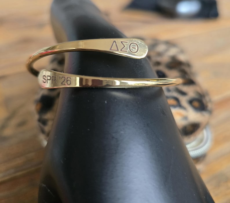 May include: A gold-colored cuff bracelet with the Greek letters &Delta;&Sigma;&Theta; and the text "SPR '26" engraved on it. The bracelet is wrapped around a black object, with a blurred background featuring a leopard print.