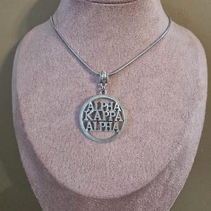 May include: Silver-toned necklace with a circular pendant. The pendant features the words "ALPHA KAPPA ALPHA" cut out within a circular frame. The necklace is displayed on a pink mannequin.