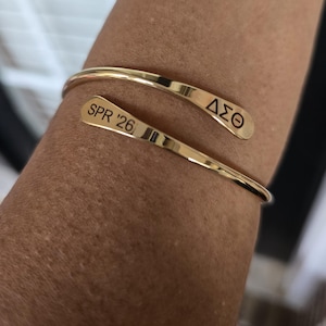 May include: A gold-colored cuff bracelet with the engraved text "SPR '26" and the Greek letters "&Delta;&Sigma;&Theta;". The bracelet is wrapped around a person's arm. The bracelet is a simple, elegant design.
