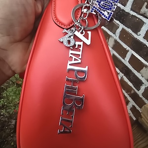 May include: A smooth, coral-red handbag featuring a silver-toned metal charm set that includes the vertical text ZETA PHI BETA, a smaller charm with the number 1920, and a blue rhinestone-encrusted charm with the letters ZOB.