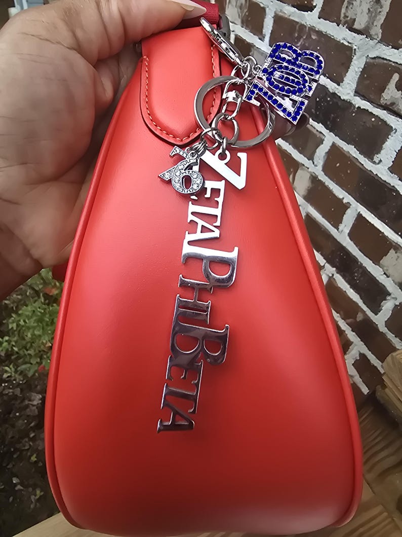 May include: A smooth, coral-red handbag featuring a silver-toned metal charm set that includes the vertical text ZETA PHI BETA, a smaller charm with the number 1920, and a blue rhinestone-encrusted charm with the letters ZOB.