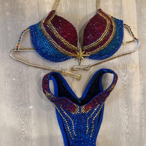 May include: A two-piece bikini set heavily embellished with red, blue, and gold crystals. The top features a gold star emblem at the center, while the bottom is a high-cut style. Both pieces are covered in intricate, sparkling rhinestone patterns.