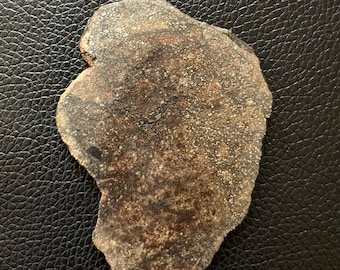 May include: A roughly-shaped, brown stone with a speckled surface. The stone has a dark, mottled appearance with lighter, granular areas. The edges are uneven, giving it a natural, organic look. The stone is set against a black, textured background.