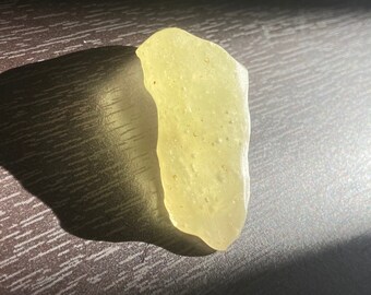 May include: A single piece of pale yellow sea glass with a translucent, frosted appearance and an irregular, weathered shape. The surface displays small, scattered indentations and a smooth, rounded texture consistent with natural erosion.